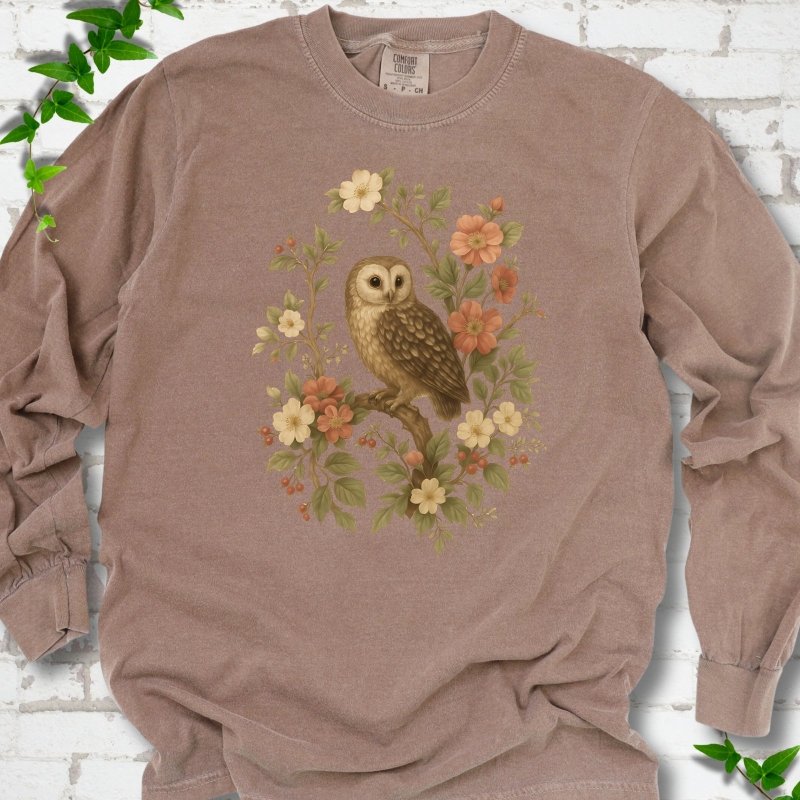 Espresso long sleeve T-shirt flat lay with a botanical illustration of a brown owl perched on a branch, framed by wild roses, cream blossoms, rosehips and olive-green leaves, on white brick with ivy.