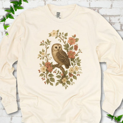Ivory long sleeve T-shirt flat lay with a botanical illustration of a brown owl perched on a branch, framed by wild roses, cream blossoms, rosehips and olive-green leaves, on white brick with ivy.