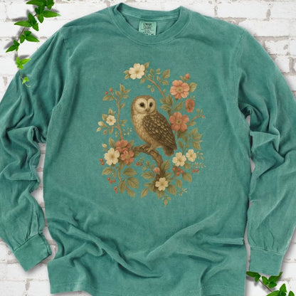 Light Green long sleeve T-shirt flat lay with a botanical illustration of a brown owl perched on a branch, framed by wild roses, cream blossoms, rosehips and olive-green leaves, on white brick with ivy.