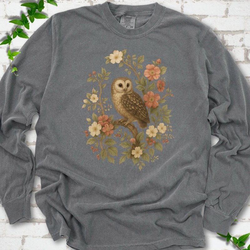 Pepper long sleeve T-shirt flat lay with a botanical illustration of a brown owl perched on a branch, framed by wild roses, cream blossoms, rosehips and olive-green leaves, on white brick with ivy.