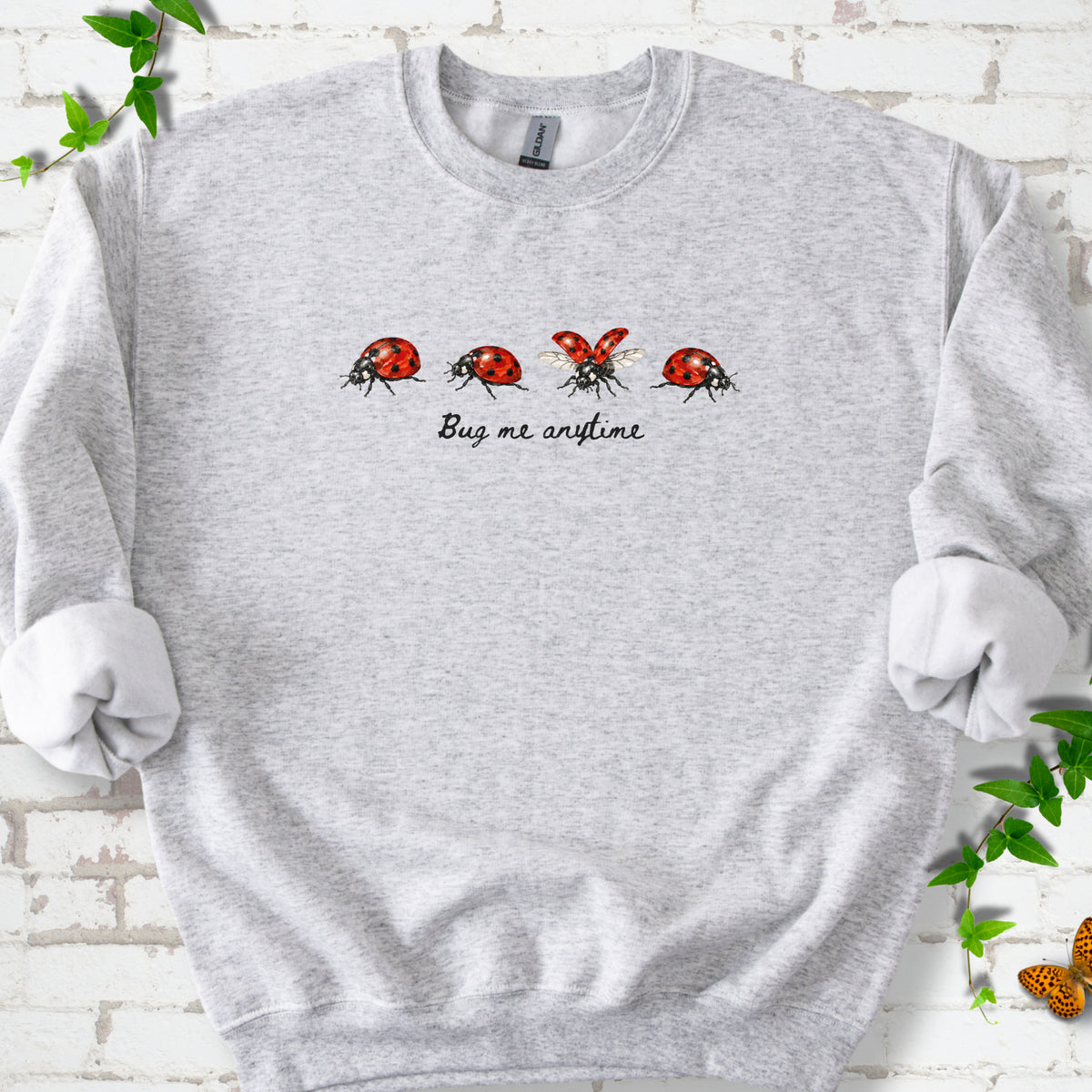 Ash classic sweatshirt laid flat, showing four red and black ladybugs above "Bug me anytime" lettering.