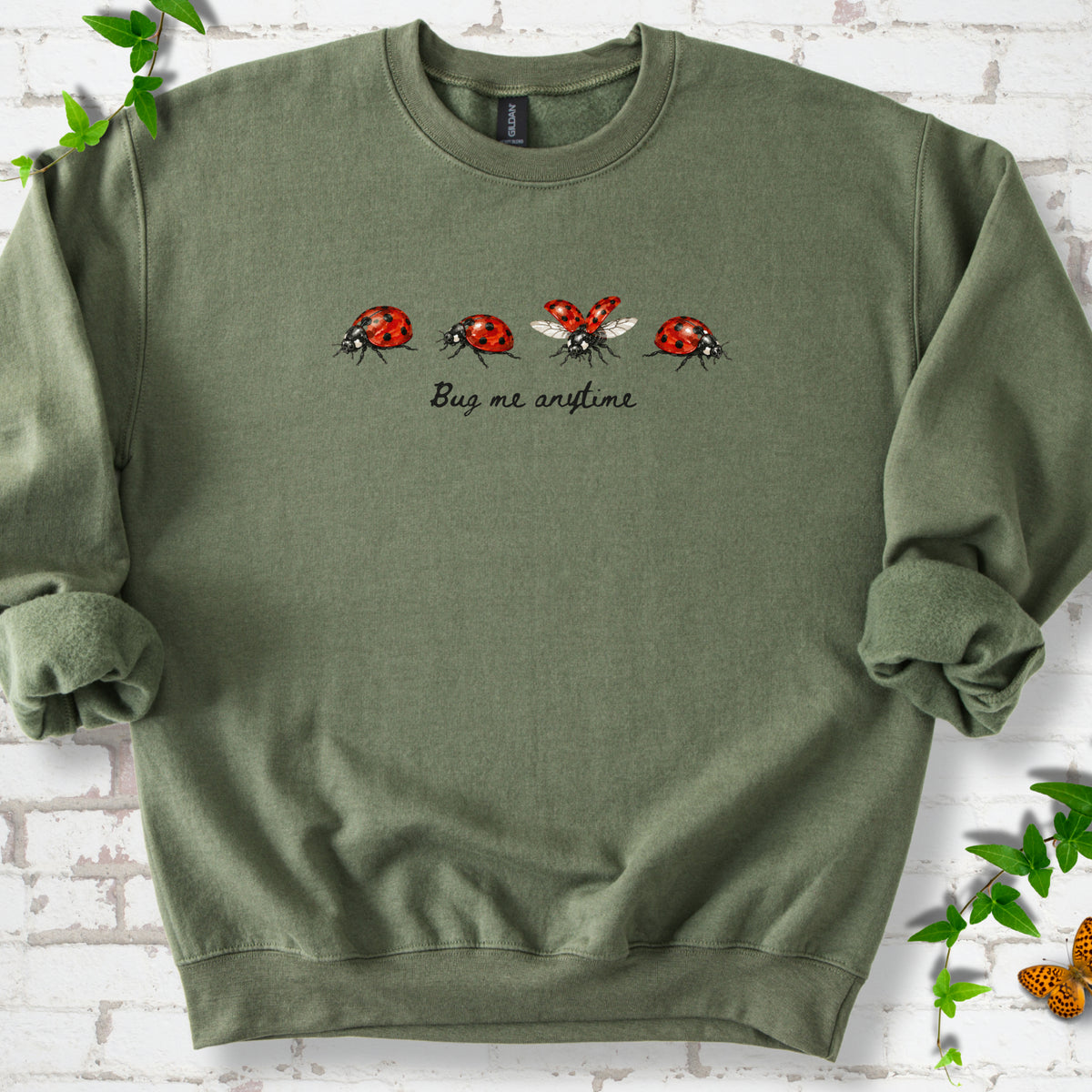 Military Green classic sweatshirt laid flat, showing four red and black ladybugs above "Bug me anytime" lettering.