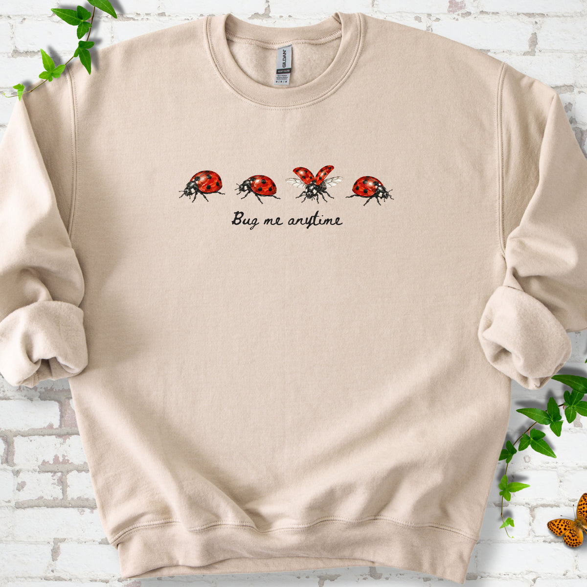 Sand classic sweatshirt laid flat, showing four red and black ladybugs above "Bug me anytime" lettering.