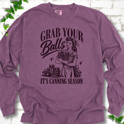 Berry long sleeve T-shirt flat lay with a dark plum retro illustration of a 1950s-style woman in a headscarf and apron hugging three filled canning jars, more jars and vegetables at her feet, with "Grab your" above, "Balls" in script and "It's canning season" below, on white brick with ivy.