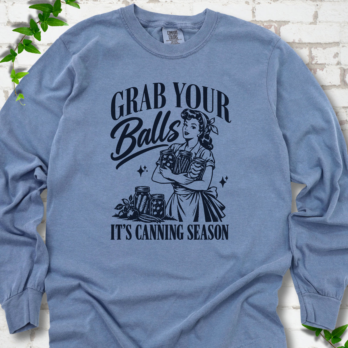 Blue Jean long sleeve T-shirt flat lay with a navy retro illustration of a 1950s-style woman in a headscarf and apron hugging three filled canning jars, more jars and vegetables at her feet, with "Grab your" above, "Balls" in script and "It's canning season" below, on white brick with ivy.