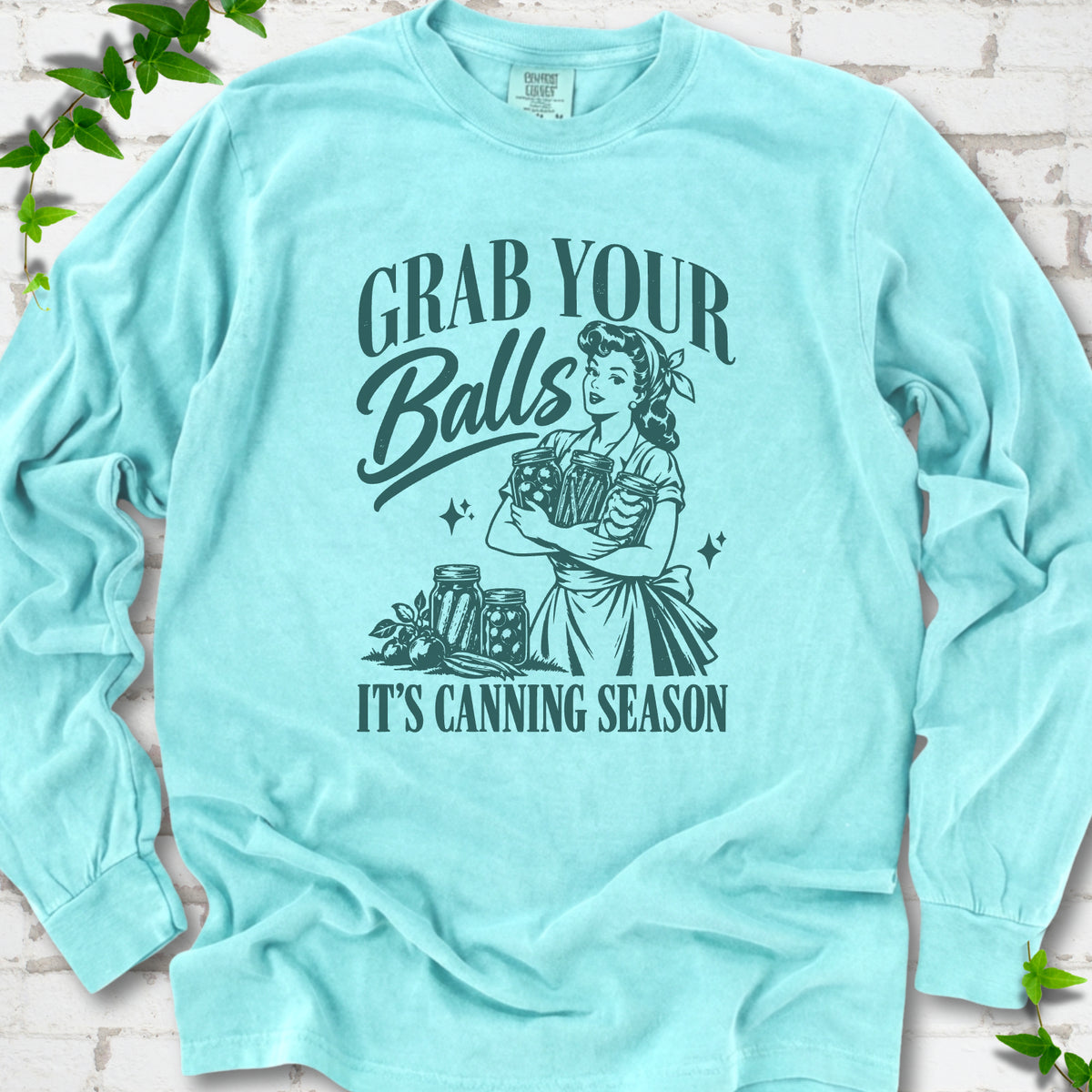 Chalky Mint long sleeve T-shirt flat lay with a deep teal retro illustration of a 1950s-style woman in a headscarf and apron hugging three filled canning jars, more jars and vegetables at her feet, with "Grab your" above, "Balls" in script and "It's canning season" below, on white brick with ivy.