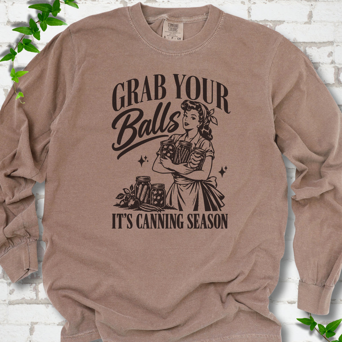 Espresso long sleeve T-shirt flat lay with a dark brown retro illustration of a 1950s-style woman in a headscarf and apron hugging three filled canning jars, more jars and vegetables at her feet, with "Grab your" above, "Balls" in script and "It's canning season" below, on white brick with ivy.
