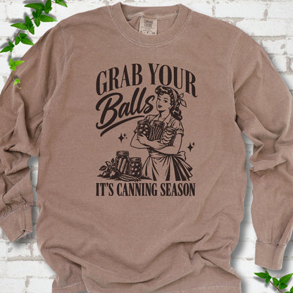 Espresso long sleeve T-shirt flat lay with a dark brown retro illustration of a 1950s-style woman in a headscarf and apron hugging three filled canning jars, more jars and vegetables at her feet, with "Grab your" above, "Balls" in script and "It's canning season" below, on white brick with ivy.