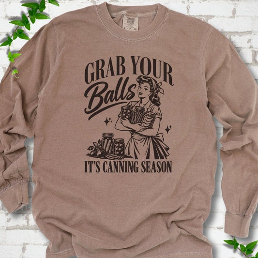 Espresso long sleeve T-shirt flat lay with a dark brown retro illustration of a 1950s-style woman in a headscarf and apron hugging three filled canning jars, more jars and vegetables at her feet, with "Grab your" above, "Balls" in script and "It's canning season" below, on white brick with ivy.