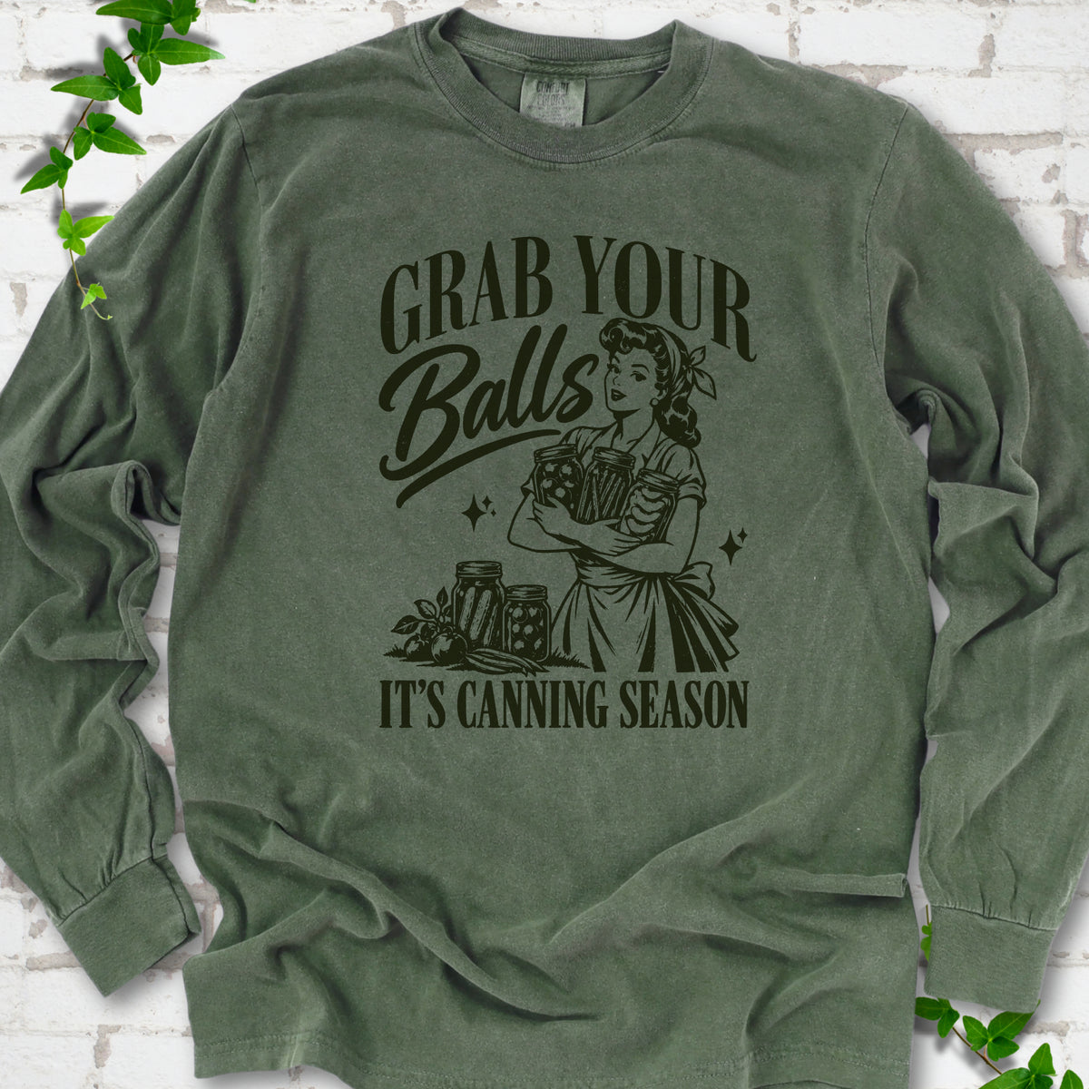 Hemp long sleeve T-shirt flat lay with a dark olive retro illustration of a 1950s-style woman in a headscarf and apron hugging three filled canning jars, more jars and vegetables at her feet, with "Grab your" above, "Balls" in script and "It's canning season" below, on white brick with ivy.