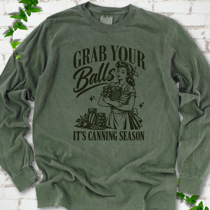Hemp long sleeve T-shirt flat lay with a dark olive retro illustration of a 1950s-style woman in a headscarf and apron hugging three filled canning jars, more jars and vegetables at her feet, with "Grab your" above, "Balls" in script and "It's canning season" below, on white brick with ivy.