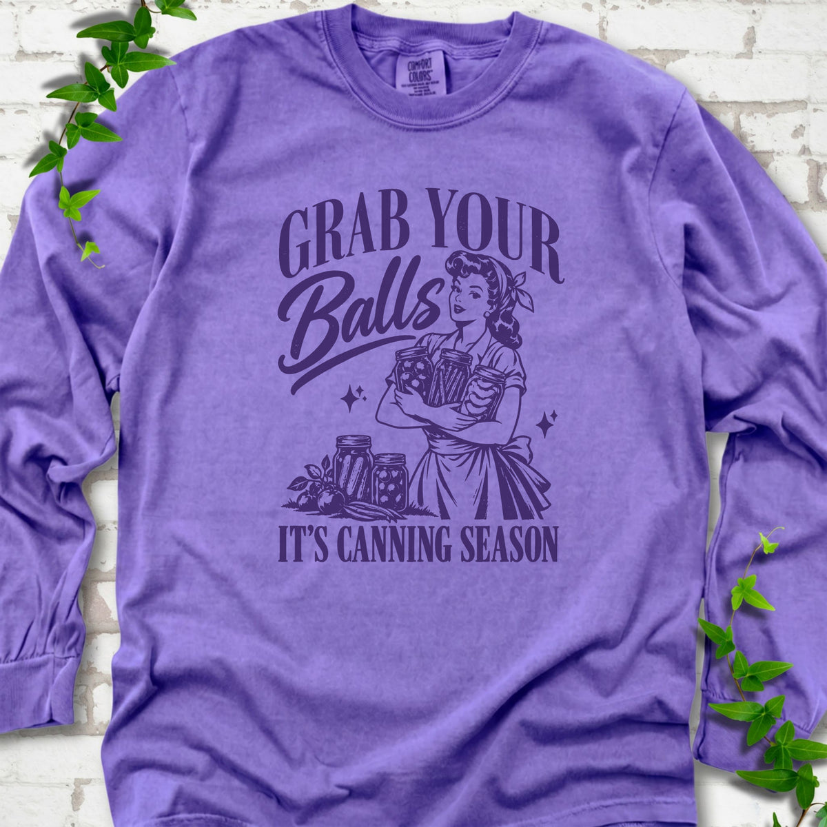 Violet long sleeve T-shirt flat lay with a deep purple retro illustration of a 1950s-style woman in a headscarf and apron hugging three filled canning jars, more jars and vegetables at her feet, with "Grab your" above, "Balls" in script and "It's canning season" below, on white brick with ivy.