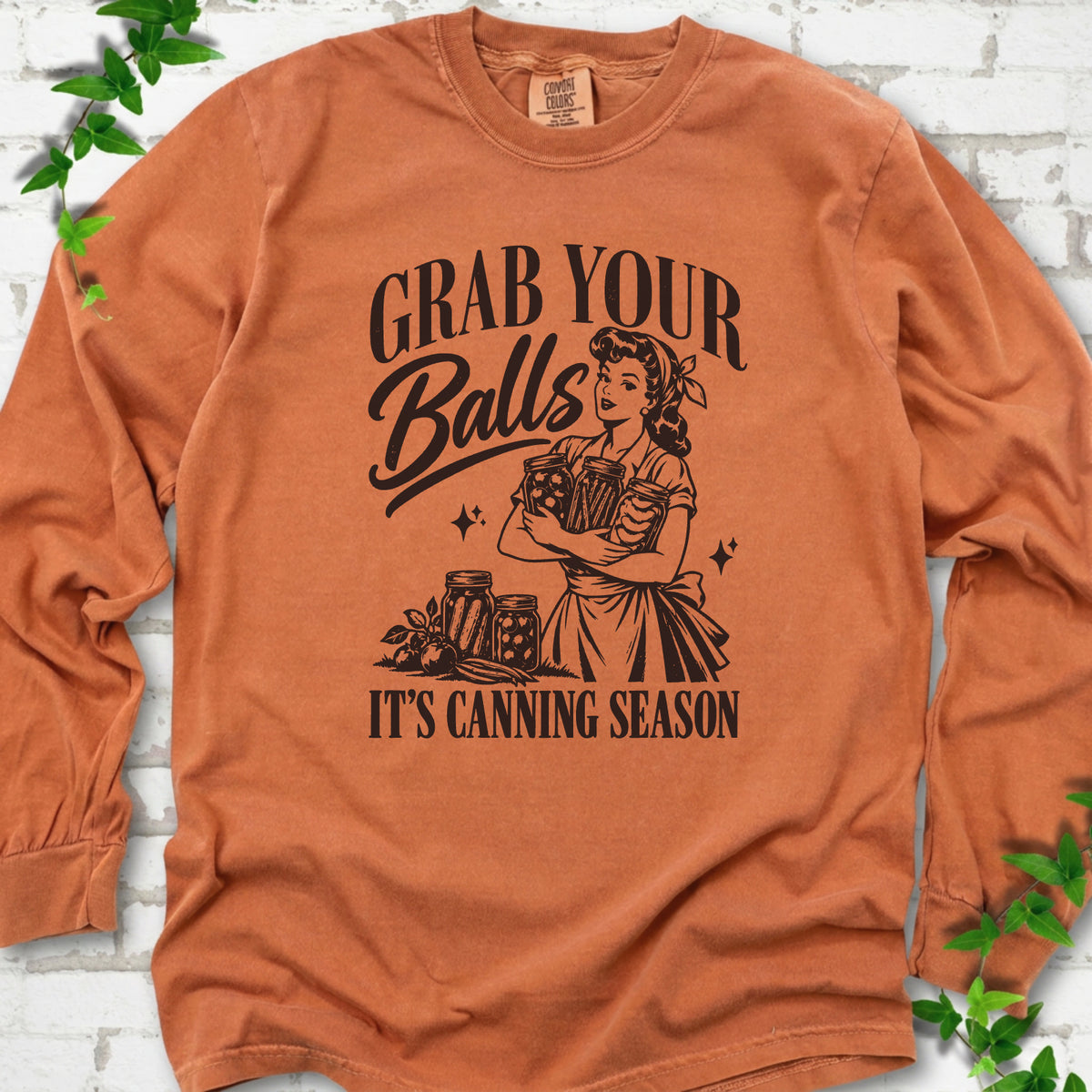 Yam long sleeve T-shirt flat lay with a dark brown retro illustration of a 1950s-style woman in a headscarf and apron hugging three filled canning jars, more jars and vegetables at her feet, with "Grab your" above, "Balls" in script and "It's canning season" below, on white brick with ivy.