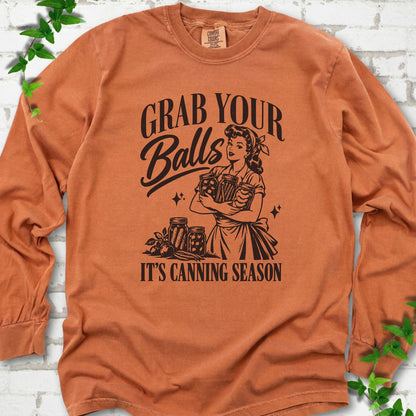Yam long sleeve T-shirt flat lay with a dark brown retro illustration of a 1950s-style woman in a headscarf and apron hugging three filled canning jars, more jars and vegetables at her feet, with "Grab your" above, "Balls" in script and "It's canning season" below, on white brick with ivy.
