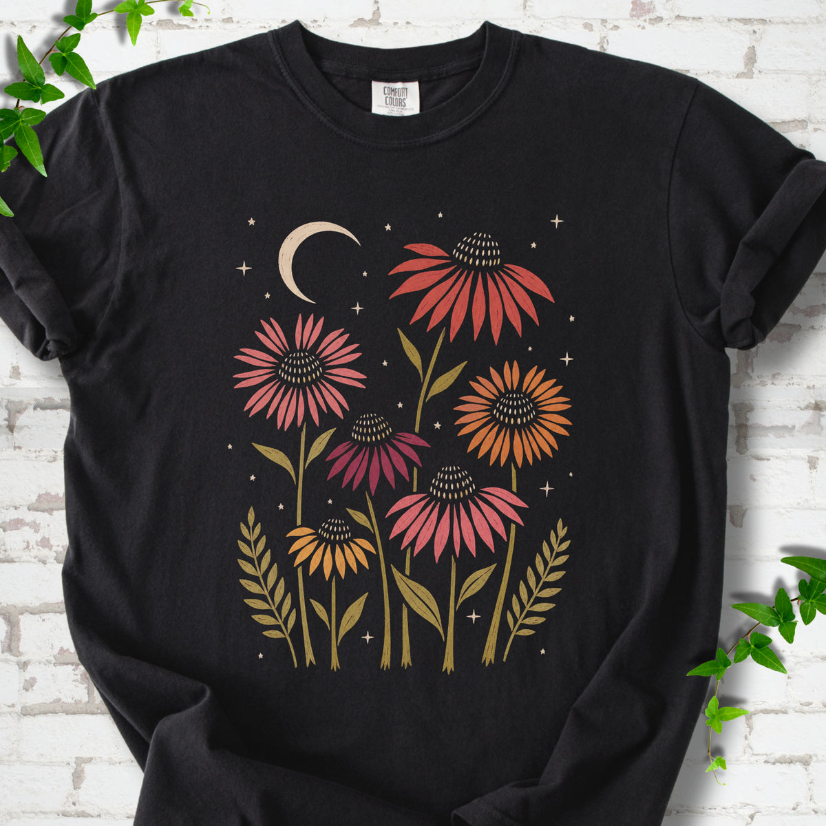 Black T-shirt laid flat, showing warm-toned coneflowers with green leaves, a cream crescent moon and small stars.