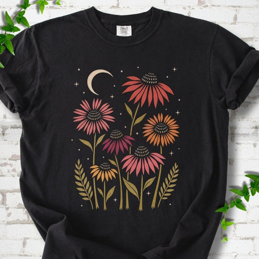 Black T-shirt laid flat, showing warm-toned coneflowers with green leaves, a cream crescent moon and small stars.