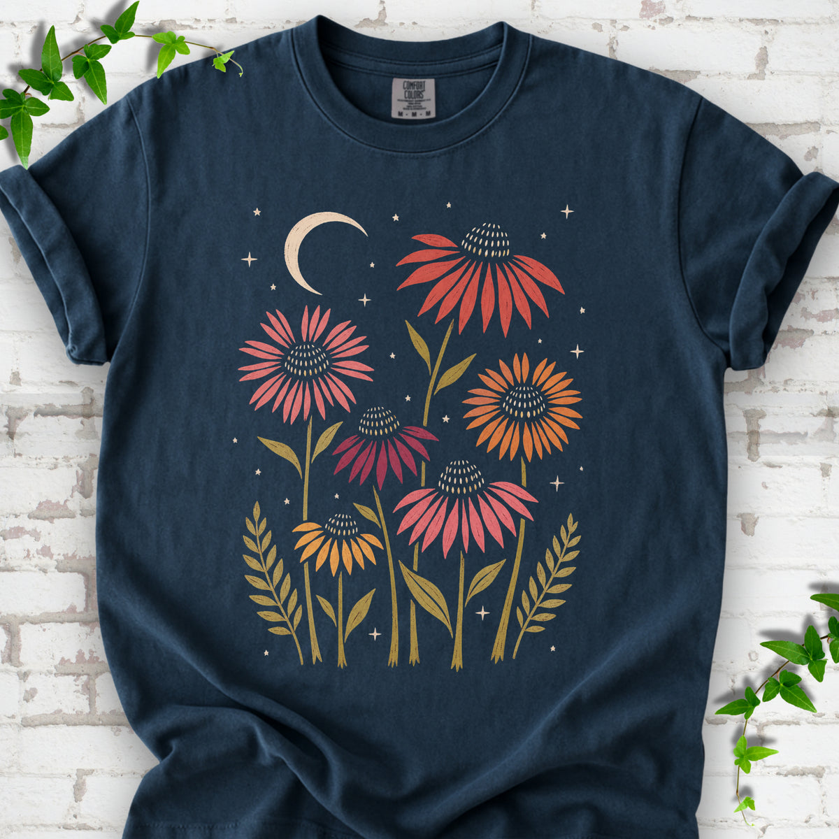 Midnight T-shirt laid flat, showing warm-toned coneflowers with green leaves, a cream crescent moon and small stars.