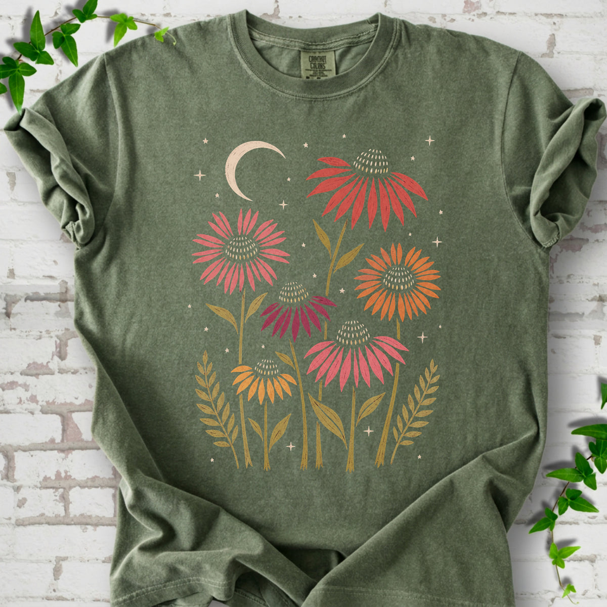 Moss T-shirt laid flat, showing warm-toned coneflowers with green leaves, a cream crescent moon and small stars.