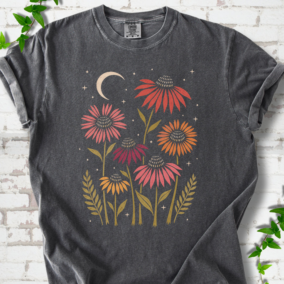 Pepper T-shirt laid flat, showing warm-toned coneflowers with green leaves, a cream crescent moon and small stars.