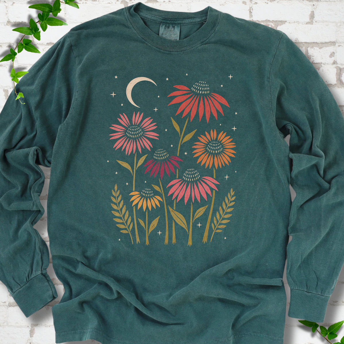 Blue Spruce long sleeve tee laid flat, showing warm-toned coneflowers with green leaves, a cream crescent moon and small stars.