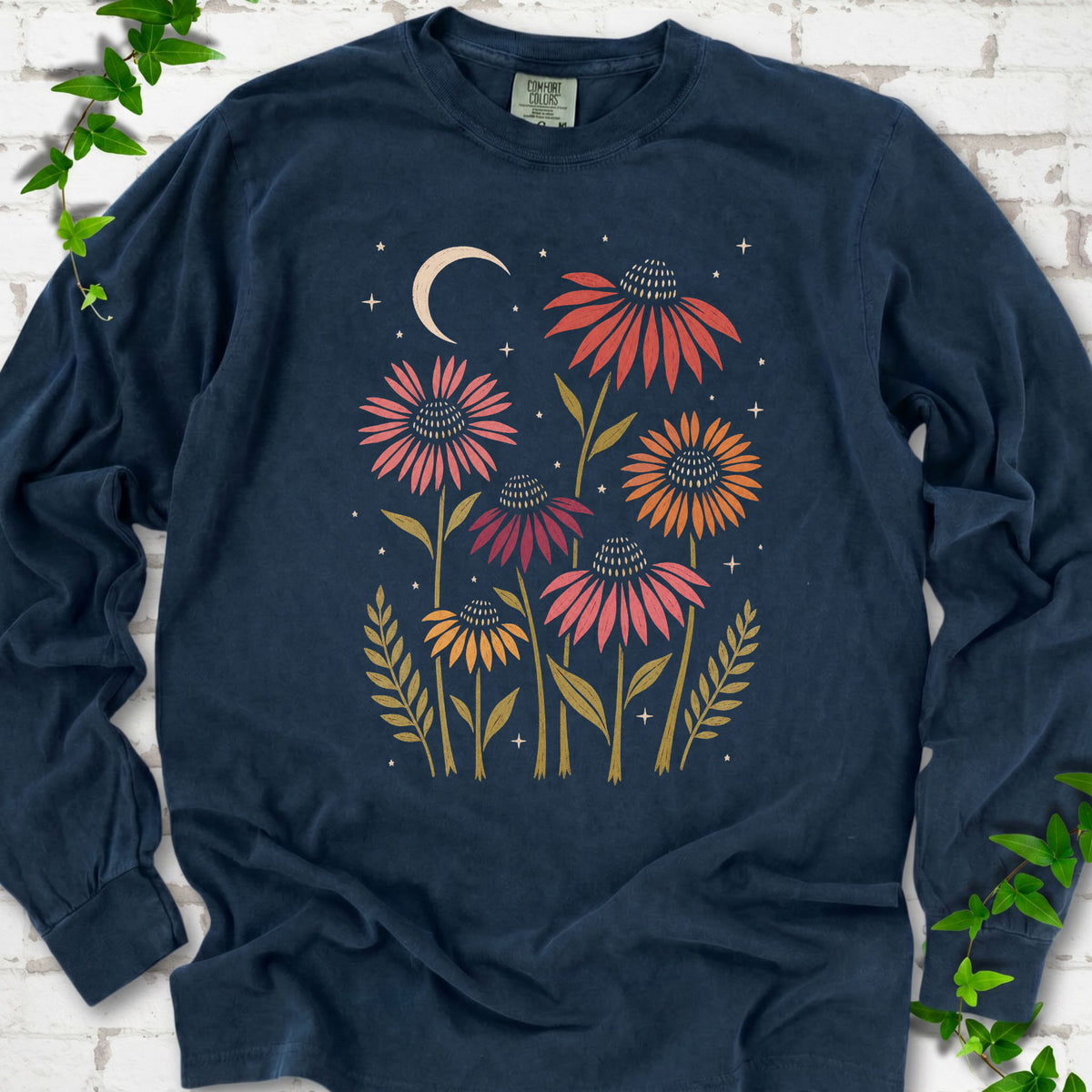 Midnight long sleeve tee laid flat, showing warm-toned coneflowers with green leaves, a cream crescent moon and small stars.