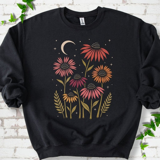 Black classic sweatshirt laid flat, showing warm-toned coneflowers with green leaves, a cream crescent moon and small stars.