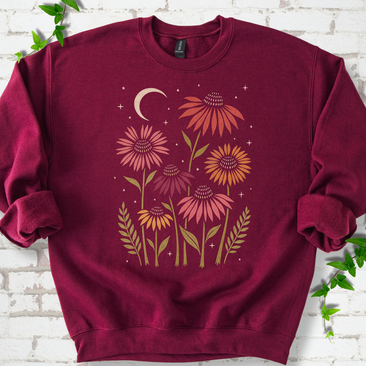Maroon classic sweatshirt laid flat, showing warm-toned coneflowers with green leaves, a cream crescent moon and small stars.