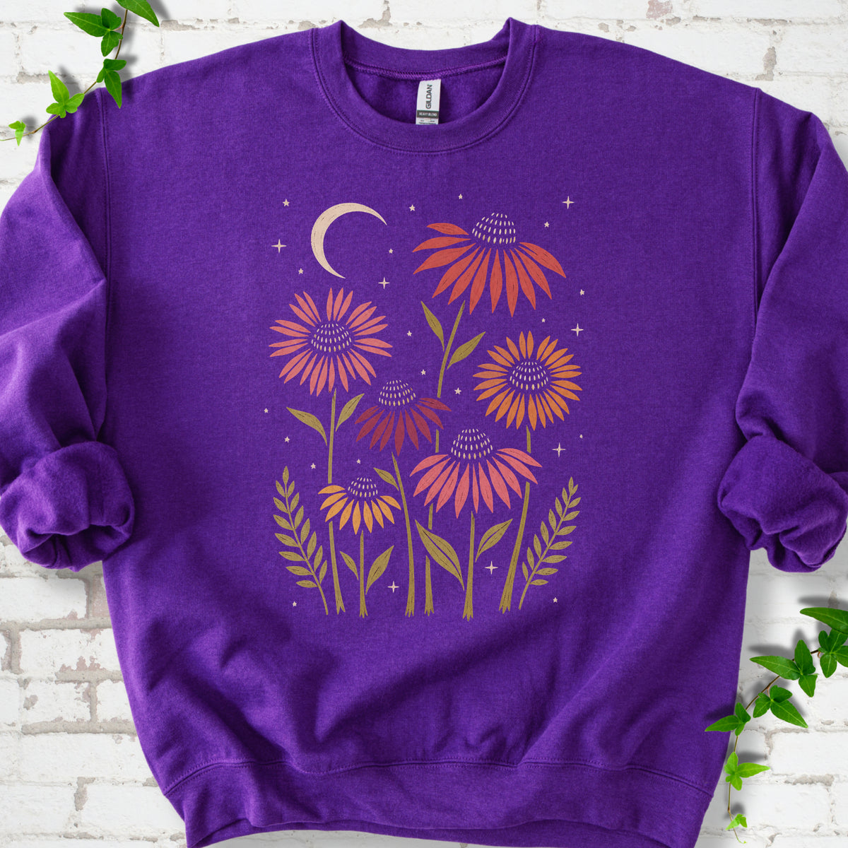 Purple classic sweatshirt laid flat, showing warm-toned coneflowers with green leaves, a cream crescent moon and small stars.