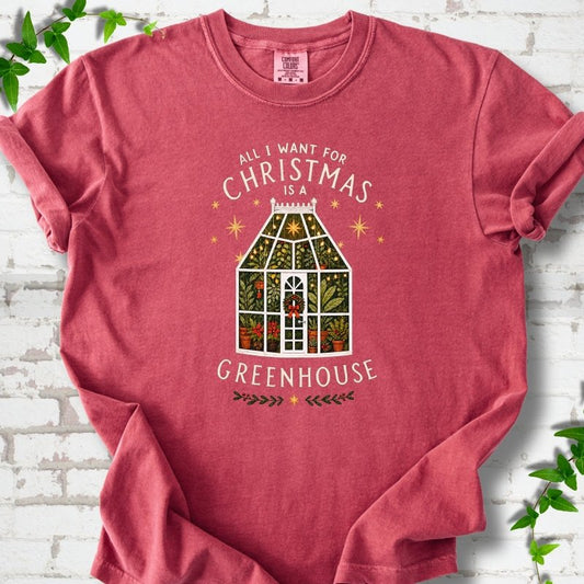 Crimson T-shirt laid flat, featuring a plant-filled glass greenhouse with a red bow, gold stars and the slogan 'All I want for Christmas is a greenhouse'.