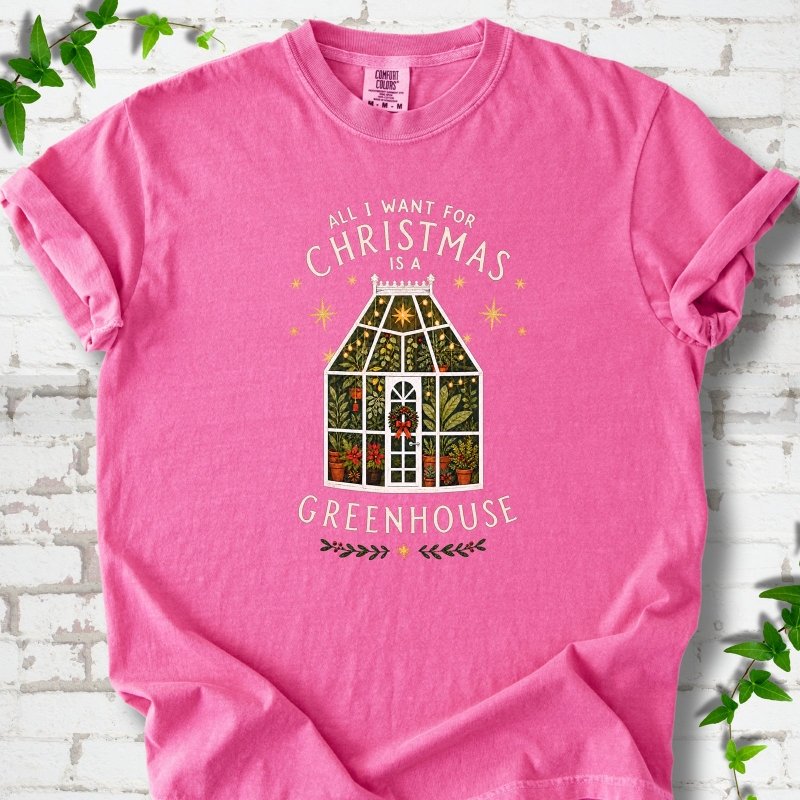 Crunchberry T-shirt laid flat, featuring a plant-filled glass greenhouse with a red bow, gold stars and the slogan 'All I want for Christmas is a greenhouse'.