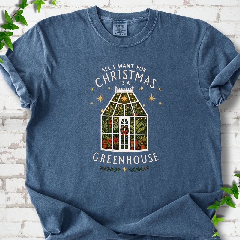 Denim T-shirt laid flat, featuring a plant-filled glass greenhouse with a red bow, gold stars and the slogan 'All I want for Christmas is a greenhouse'.