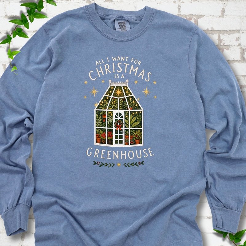 Blue Jean long sleeve T-shirt laid flat, featuring a plant-filled glass greenhouse with a red bow, gold stars and the slogan 'All I want for Christmas is a greenhouse'.