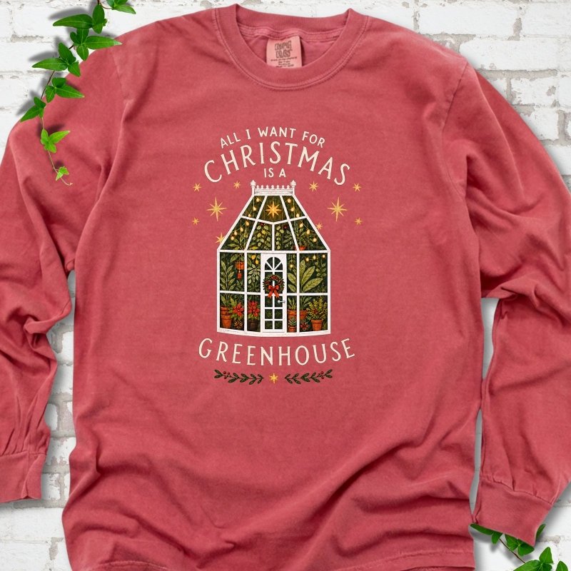 Crimson long sleeve T-shirt laid flat, featuring a plant-filled glass greenhouse with a red bow, gold stars and the slogan 'All I want for Christmas is a greenhouse'.