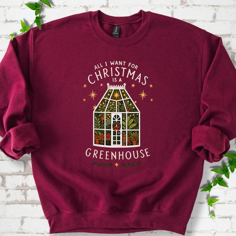 Maroon sweatshirt laid flat, featuring a plant-filled glass greenhouse with a red bow, gold stars and the slogan 'All I want for Christmas is a greenhouse'.