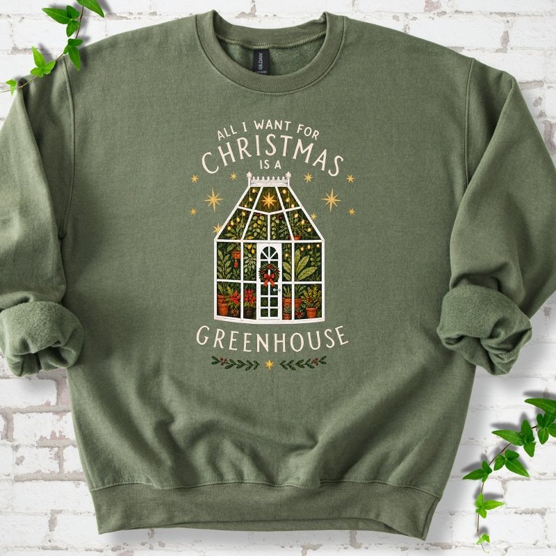Military Green sweatshirt laid flat, featuring a plant-filled glass greenhouse with a red bow, gold stars and the slogan 'All I want for Christmas is a greenhouse'.