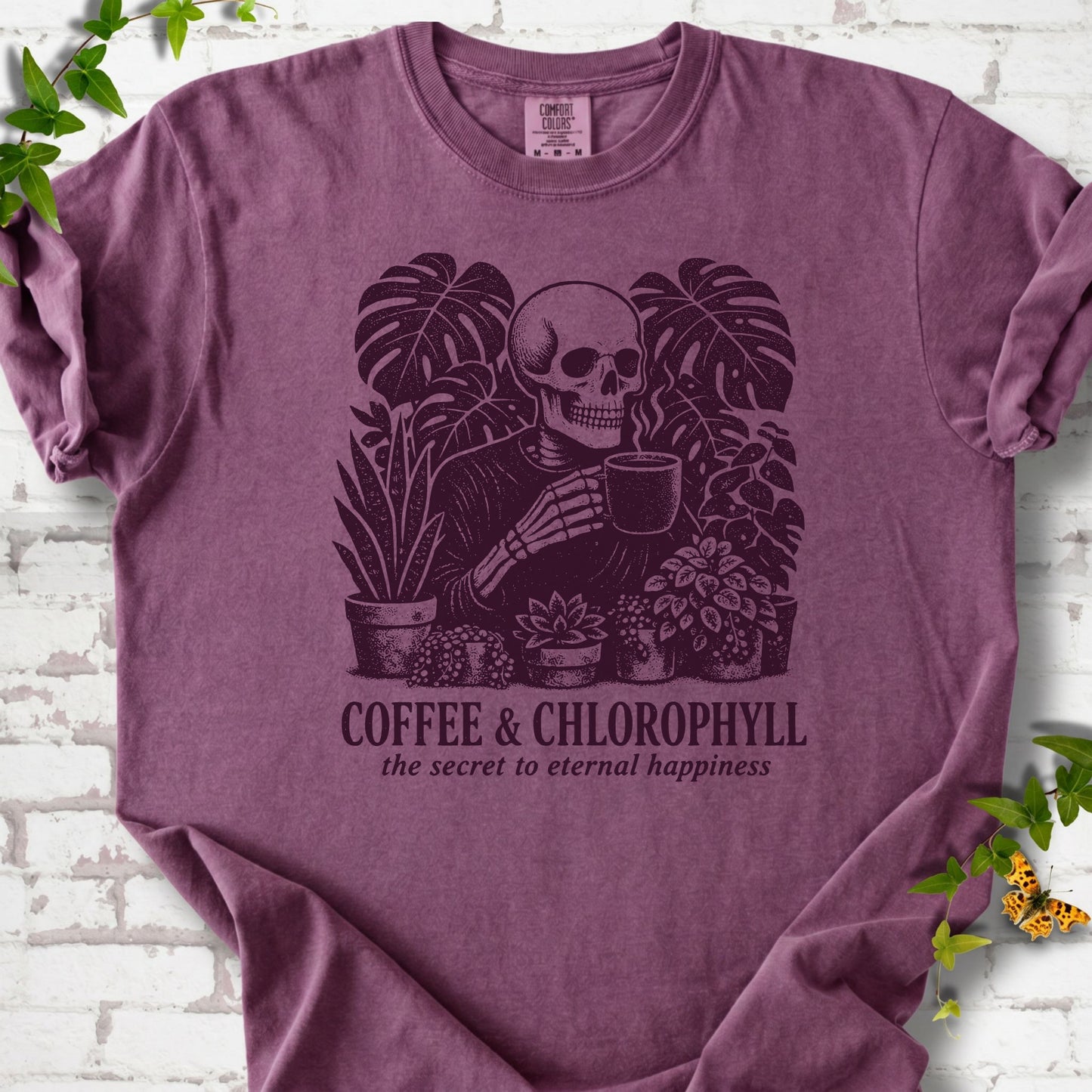 Berry Comfort Colors® short sleeve T-shirt flat lay featuring a distressed skeleton drinking coffee among houseplants