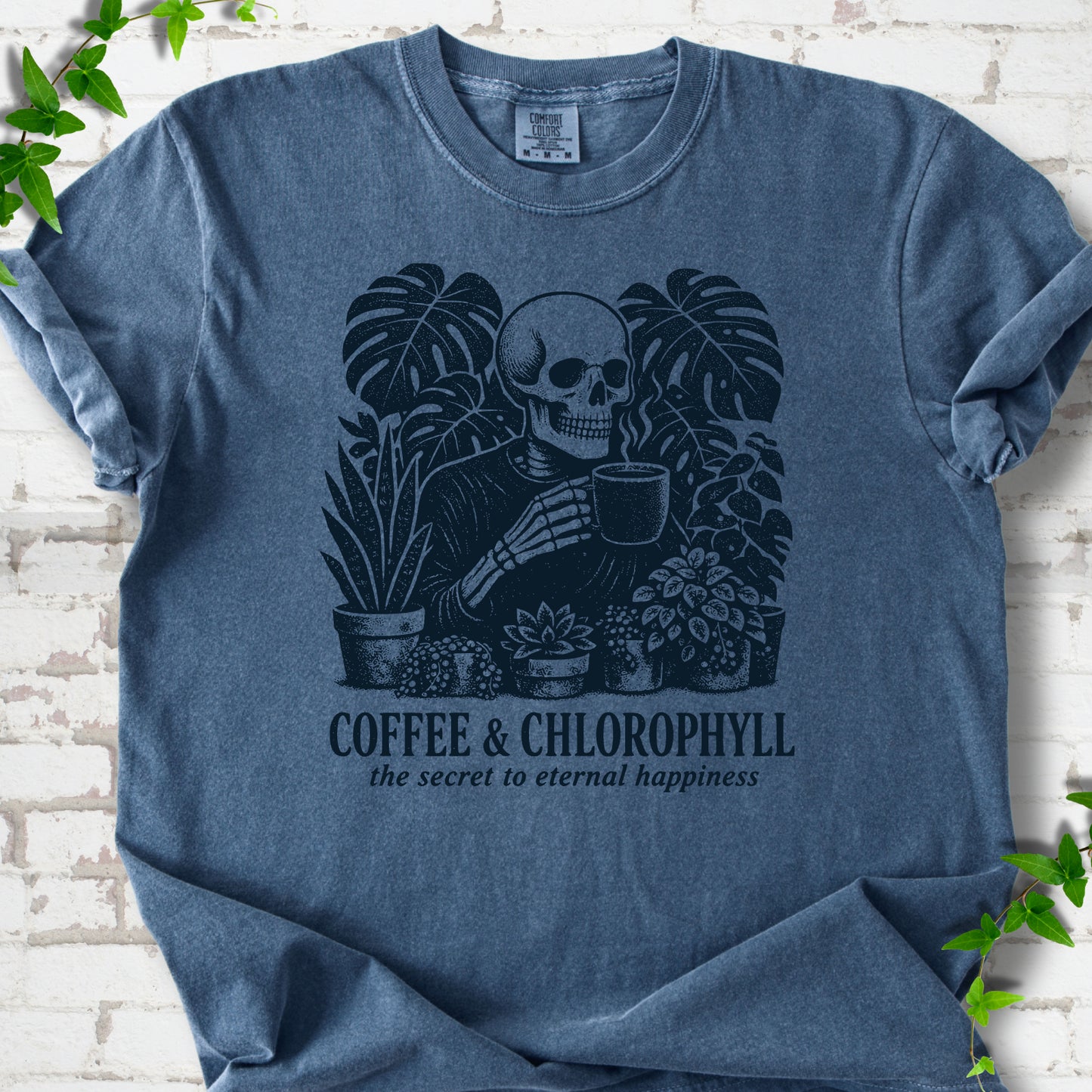 Denim Comfort Colors® short sleeve T-shirt flat lay featuring a distressed skeleton drinking coffee among houseplants