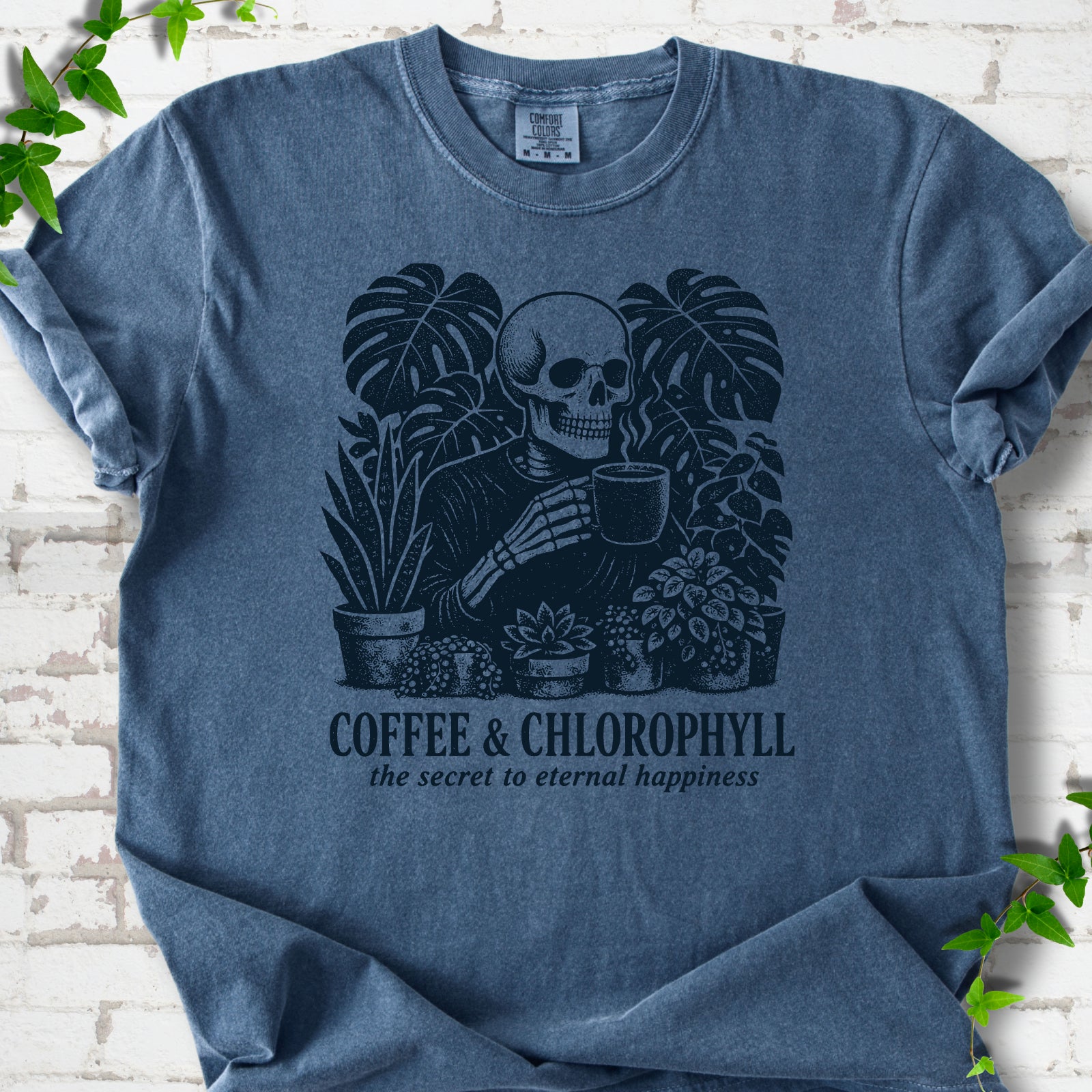 Denim Comfort Colors® short sleeve T-shirt flat lay featuring a distressed skeleton drinking coffee among houseplants