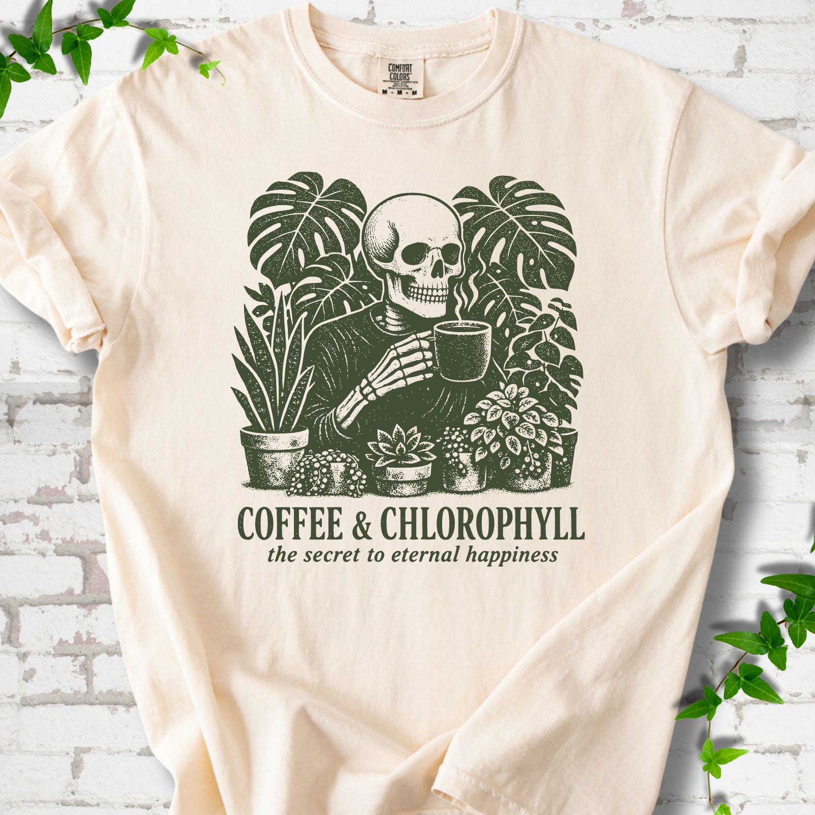 Ivory Comfort Colors® short sleeve T-shirt flat lay featuring a distressed skeleton drinking coffee among houseplants