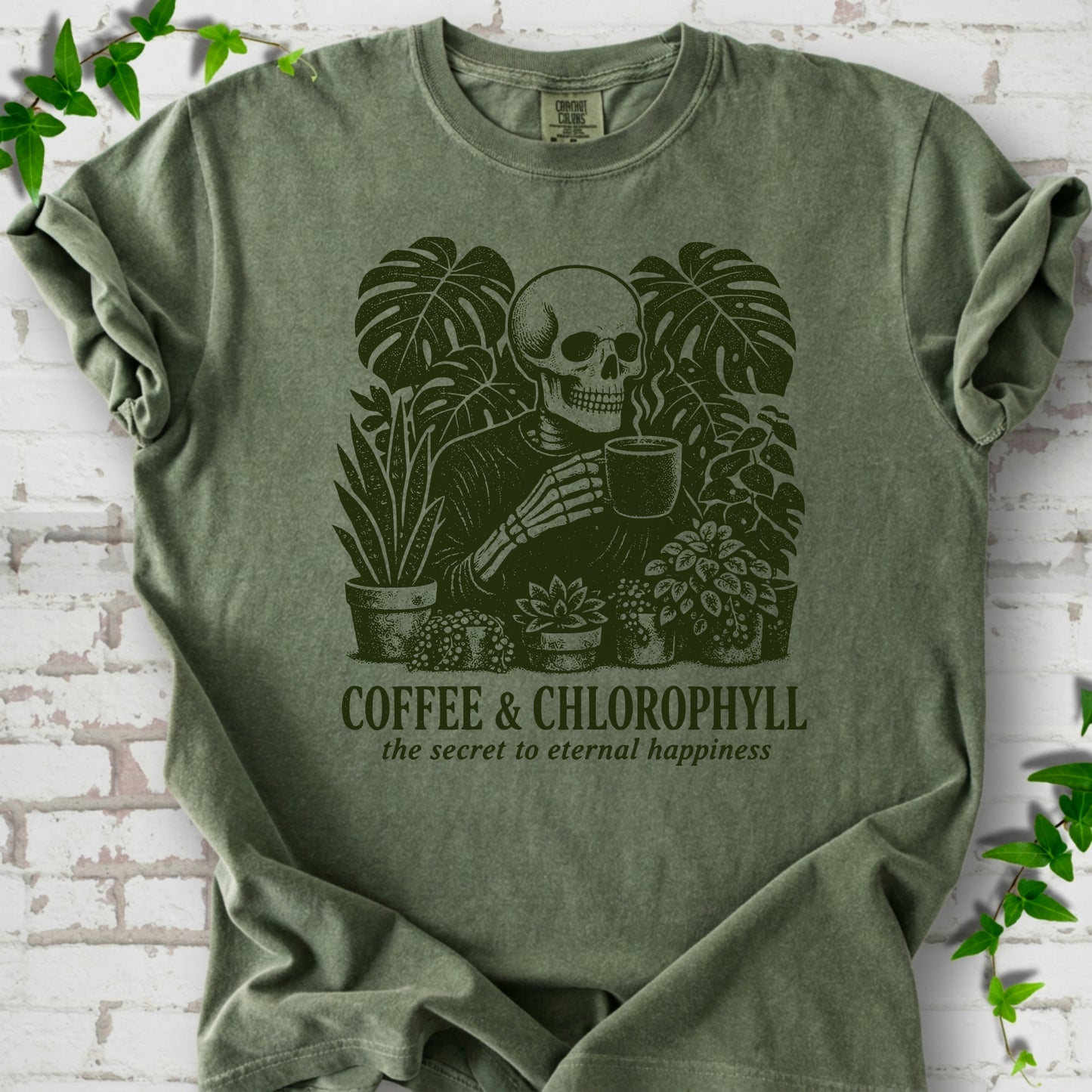 Moss Comfort Colors® short sleeve T-shirt flat lay featuring a distressed skeleton drinking coffee among houseplants