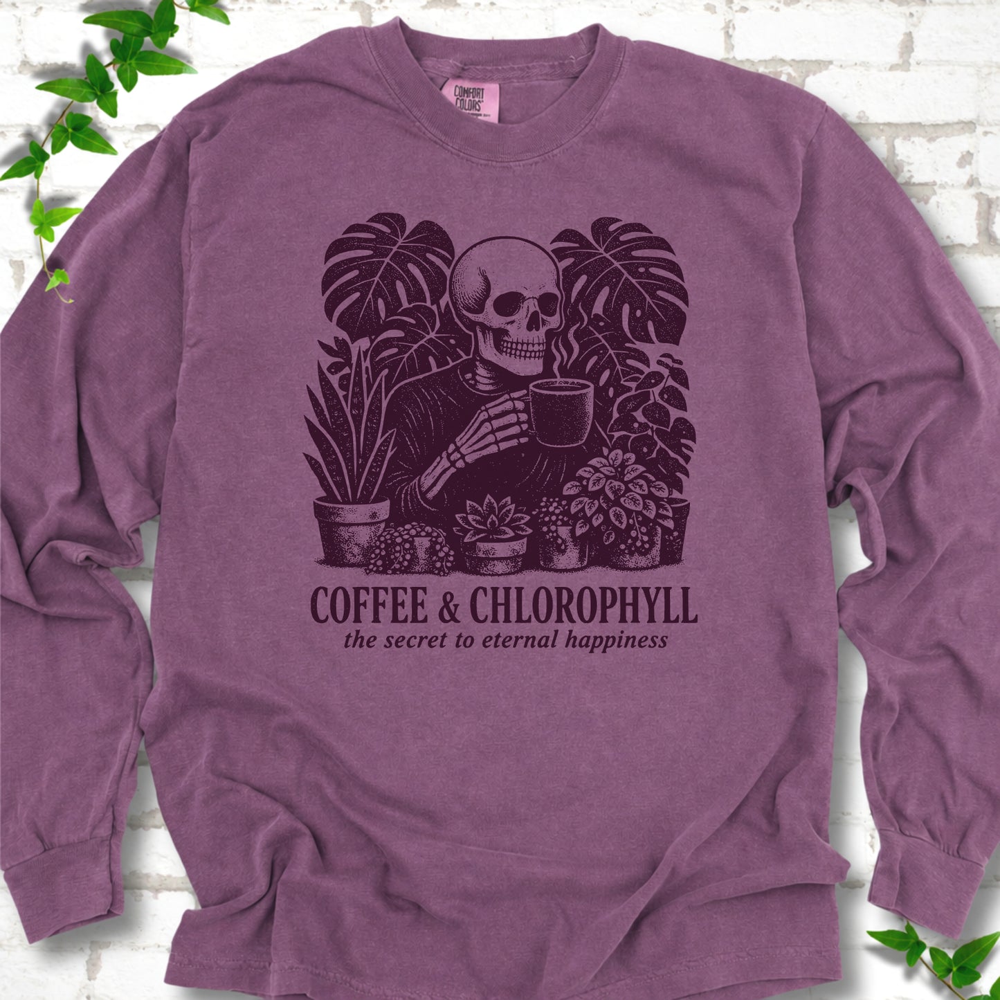 Berry Comfort Colors® long sleeve T-shirt flat lay featuring a distressed skeleton drinking coffee among houseplants