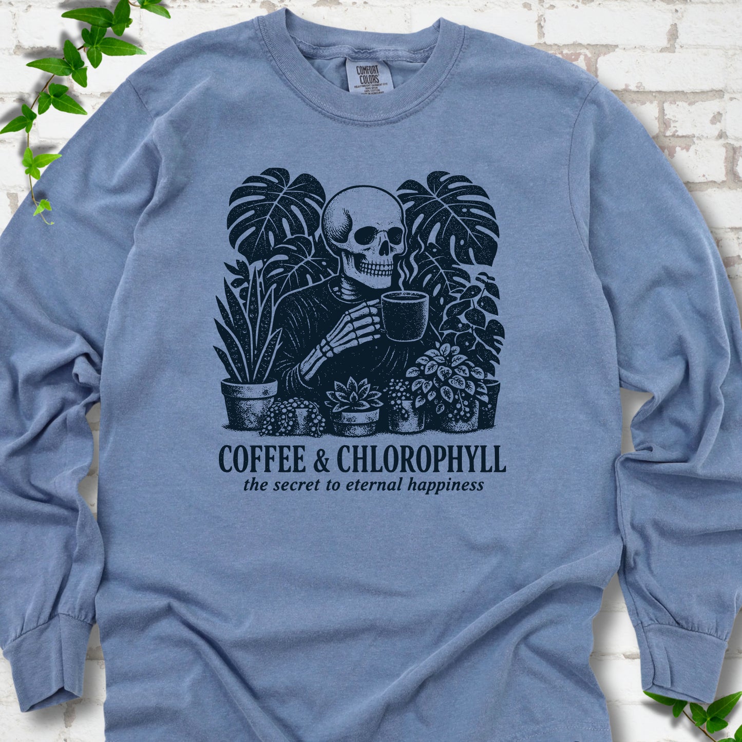 Blue Jean Comfort Colors® long sleeve T-shirt flat lay featuring a distressed skeleton drinking coffee among houseplants