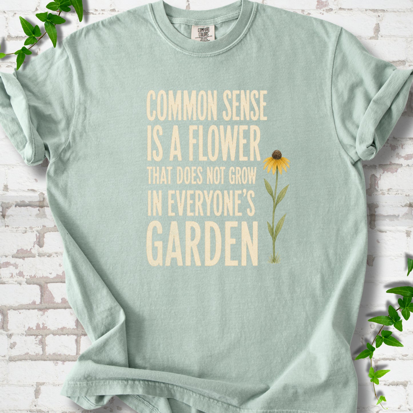Bay Comfort Colors® short sleeve T-shirt flat lay with cream text reading "Common sense is a flower that does not grow in everyone's garden" beside a yellow flower