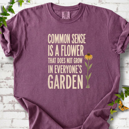 Berry Comfort Colors® short sleeve T-shirt flat lay with cream text reading "Common sense is a flower that does not grow in everyone's garden" beside a yellow flower