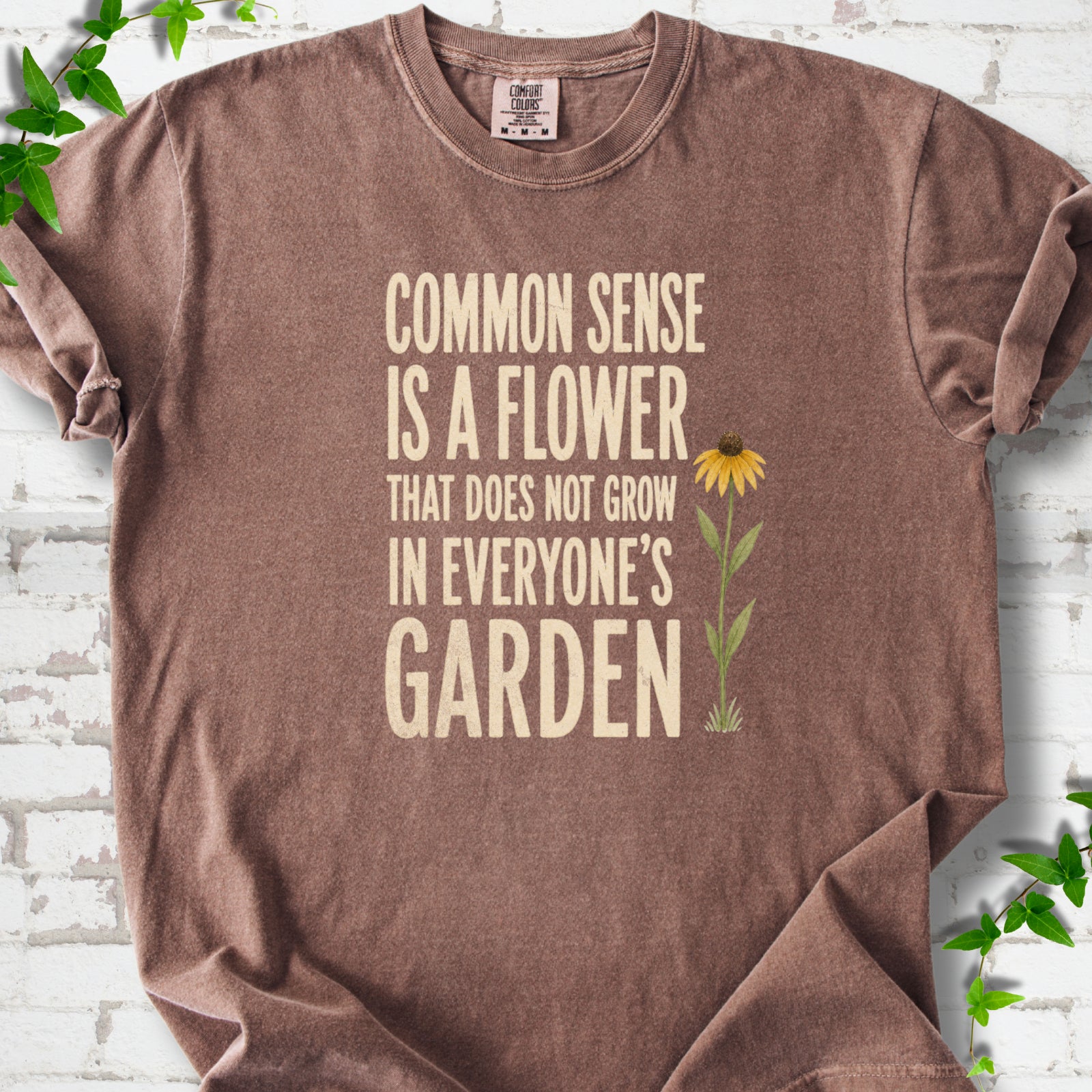 Espresso Comfort Colors® short sleeve T-shirt flat lay with cream text reading "Common sense is a flower that does not grow in everyone's garden" beside a yellow flower