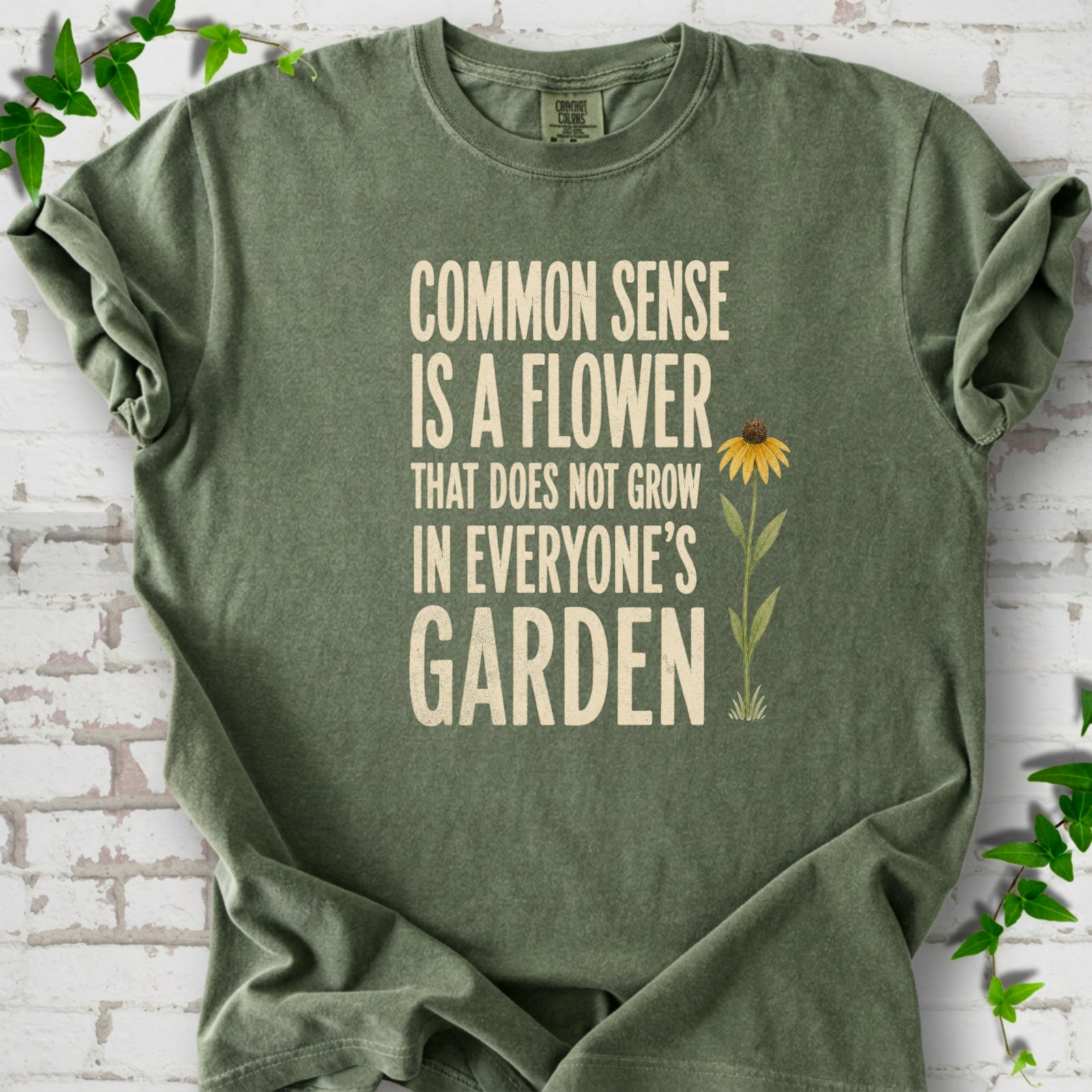 Moss Comfort Colors® short sleeve T-shirt flat lay with cream text reading "Common sense is a flower that does not grow in everyone's garden" beside a yellow flower