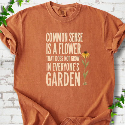 Yam Comfort Colors® short sleeve T-shirt flat lay with cream text reading "Common sense is a flower that does not grow in everyone's garden" beside a yellow flower