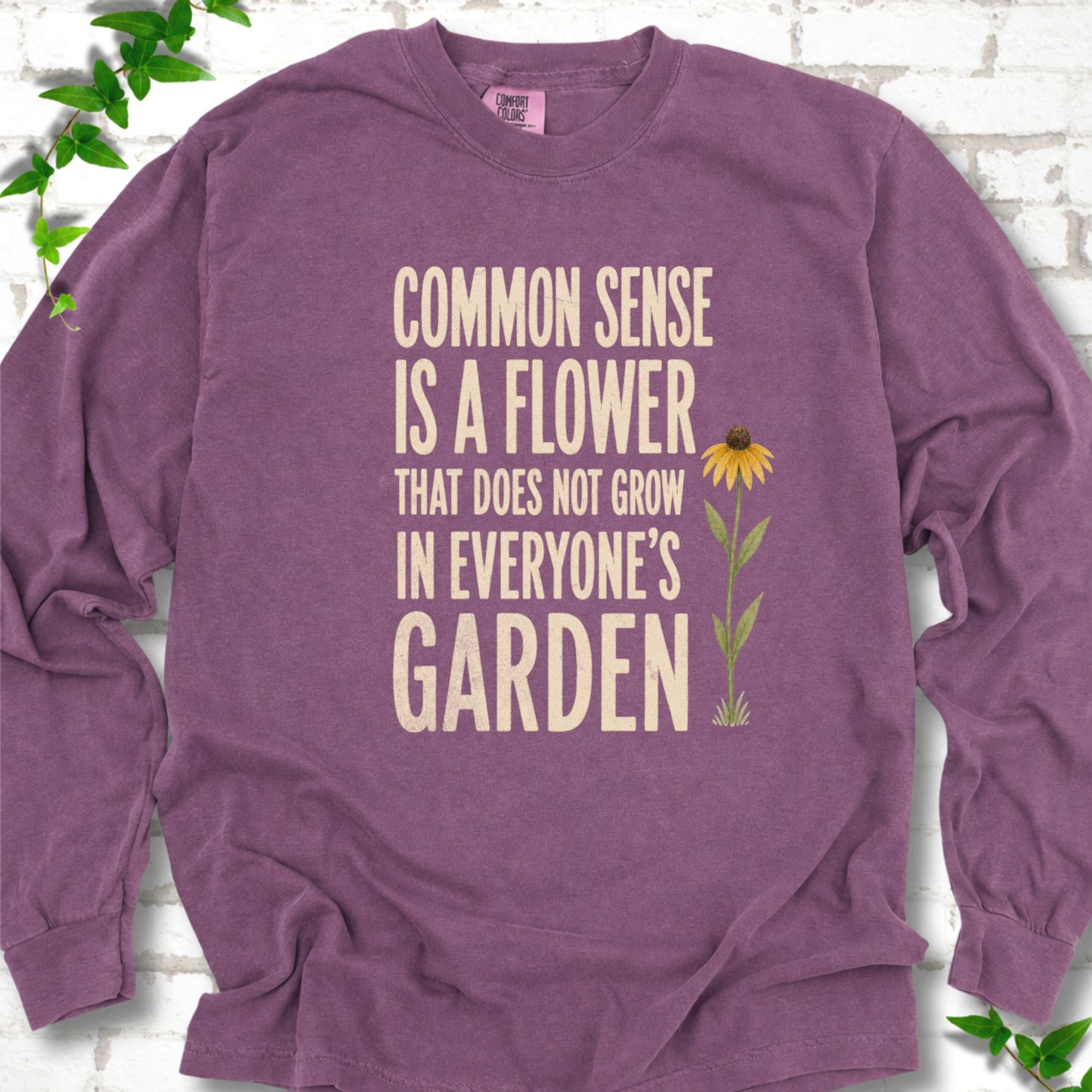 Berry Comfort Colors® long sleeve T-shirt flat lay with cream text reading "Common sense is a flower that does not grow in everyone's garden" beside a yellow flower