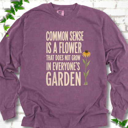 Berry Comfort Colors® long sleeve T-shirt flat lay with cream text reading "Common sense is a flower that does not grow in everyone's garden" beside a yellow flower