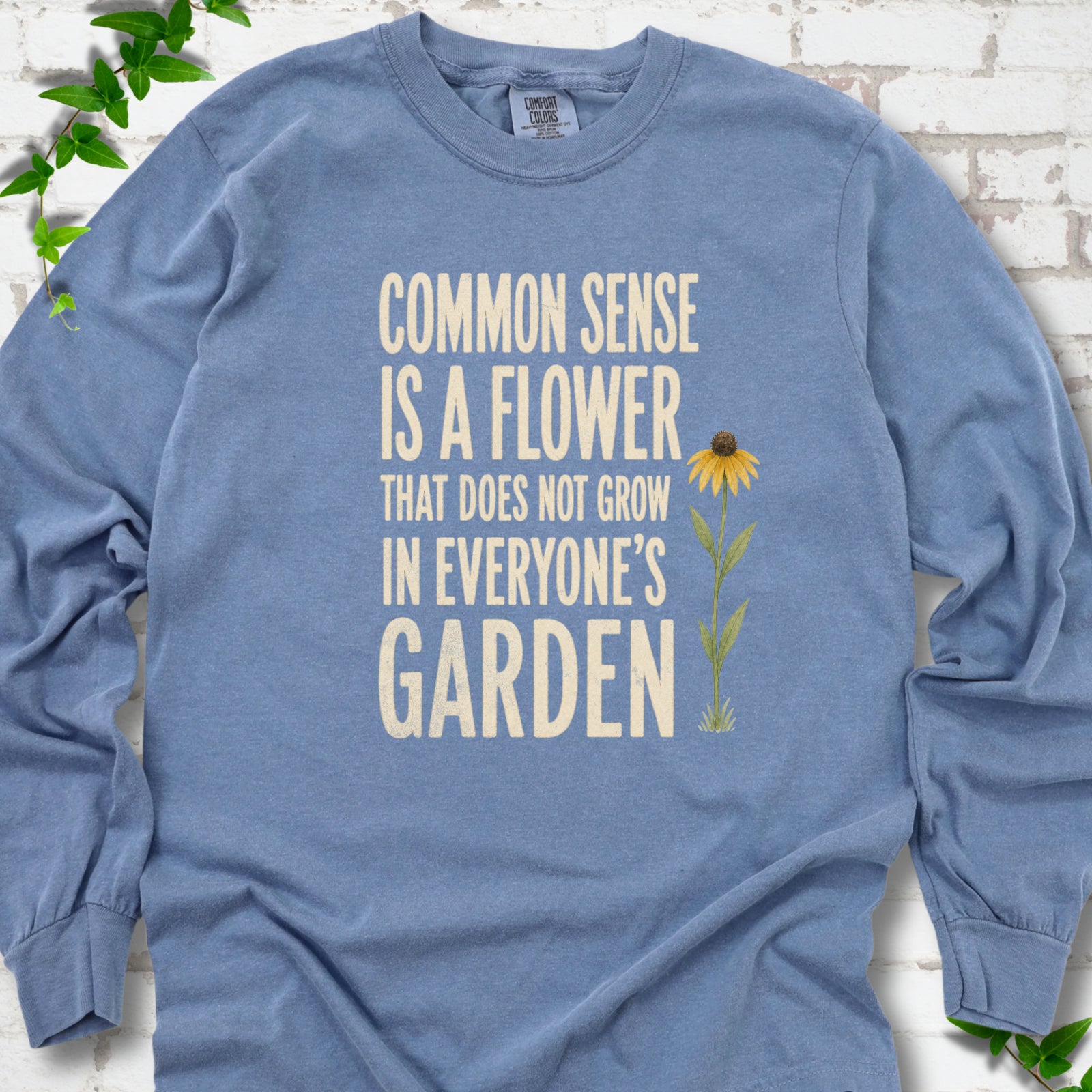 Blue Jean Comfort Colors® long sleeve T-shirt flat lay with cream text reading "Common sense is a flower that does not grow in everyone's garden" beside a yellow flower